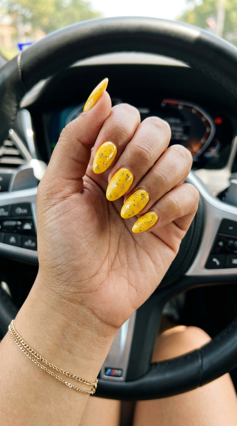 Vibrant sunny yellow almond nails with scattered metallic gold foil flakes for a playful summer vacation look. Summer nails yellow 2026.